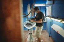 Вид сбоку квалифицированного рабочего в защитной маске с белой доской для серфинга в мастерской с голубыми стенами — стоковое фото