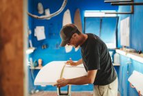 Vista lateral del hombre adulto midiendo tabla blanca mientras trabaja en un pequeño taller con paredes azules y haciendo tabla de surf - foto de stock