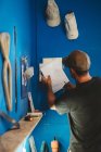 Обратный вид мастера, держащего в руках чертежи доски для серфинга, стоя в мастерской рядом с синей стеной с инструментами — стоковое фото
