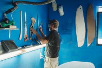 Вид збоку майстра в захисній масці, який бере інструмент з синьої стіни, працюючи в майстерні і виробляючи дошки для серфінгу — стокове фото