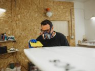 Erwachsener Handwerker in Schutzmaske und Handschuhen poliert weißes Surfbrett bei der Arbeit in kleiner Werkstatt — Stockfoto