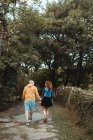 Back view of hipster couple spending happy time together while holding hands and walking on stone path among green trees — Stock Photo