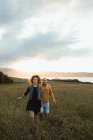 Веселая романтическая молодая пара, держащаяся за руки и идущая по зеленому полю на берегу моря на закате солнца с облачным небом — стоковое фото