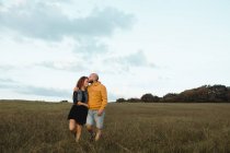 Alegre pareja joven romántica cogida de la mano y caminando en el campo verde a la orilla del mar al atardecer con cielo nublado - foto de stock