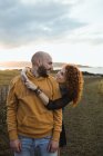 Uomo abbracciato da felice ragazza dai capelli ricci in abito e gilet di jeans mentre in piedi insieme sul prato — Foto stock