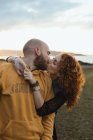 Homem abraçando por feliz cabelo encaracolado namorada no vestido e denim colete enquanto de pé juntos no prado, beijando — Fotografia de Stock