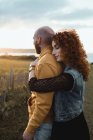 Man embracing by happy curly haired girlfriend in dress and denim vest while standing together on meadow — Stock Photo