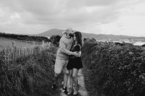 Amour jeune homme en sweat à capuche jaune embrassant petite amie en robe et gilet en denim tout en se tenant près de la clôture ensemble sur la prairie — Photo de stock