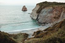 Спокійний мальовничий пейзаж з скелястими скелями, покритими зеленою травою і спокійним морем з самотньою скелею серед води в похмурий день — стокове фото