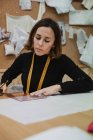 Mujer adulta enfocada sentada en la mesa y haciendo recortes mientras trabaja en un estudio de costura profesional - foto de stock
