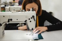 Mujer adulta sentada en la mesa y haciendo parte de la prenda en la máquina de coser mientras trabaja en un estudio profesional - foto de stock