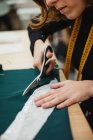 Женщина с помощью острых ножниц вырезает детали одежды из ткани, сидя за столом в швейной мастерской — стоковое фото