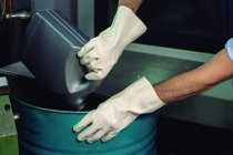 Guante manos de capataz irreconocible verter líquido químico del recipiente en el barril de metal en la fábrica - foto de stock