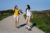 Brune lumineuse chevauchant longue planche et tenant la main de la petite amie tout en marchant sur la route dans la campagne en s'amusant — Photo de stock