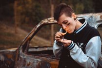 Jeune femme aux cheveux courts et peint cigarette éclairage visage avec briquet tout en se tenant près de vieille voiture rouillée dans la campagne — Photo de stock