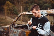 Young female with short hair and painted face lighting cigarette with lighter while standing near old rusty car in countryside — Stock Photo
