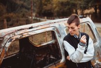 Стильна жінка з коротким волоссям курить сигарету, відпочиваючи біля старого спаленого автомобіля в сільській місцевості — стокове фото