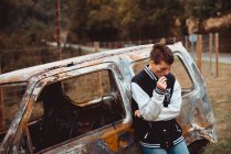 Жінка-бунтарка з коротким волоссям курить сигарету біля старого спаленого автомобіля в сільській місцевості — стокове фото