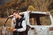 Стильный сорванец с короткими волосами и раскрашенным лицом, курящим сигарету, опирающийся на старый ржавый автомобиль в сельской местности — стоковое фото