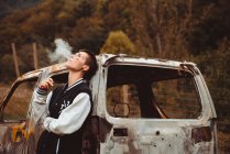 Stylish tomboy with short hair and painted face smoking cigarette as leaning on old rusty car in countryside — Stock Photo