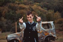 Jovem rebelde com cabelo curto e rosto pintado sorrindo e mostrando os dedos do meio enquanto estava de pé contra o carro queimado — Fotografia de Stock
