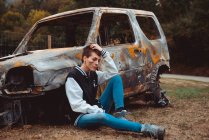 Jeune femme en tenue décontractée touchant les cheveux courts et regardant loin tout en étant assis sur le sol près de vieille voiture brûlée dans la campagne — Photo de stock