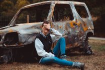 Joven hembra en traje casual tocando y escondiendo la cara mientras está sentado en el suelo cerca de viejo coche quemado en el campo - foto de stock
