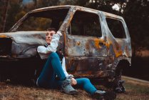 Jovem fêmea em roupa casual tocando cabelo curto e olhando para longe, sentado no chão perto de carro velho queimado no campo — Fotografia de Stock