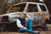 Young female in casual outfit touching short hair and looking away while sitting on ground near old burnt car in countryside — Stock Photo
