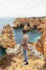 Vista lateral del tipo anónimo parado en el borde de un acantilado áspero y admirando el mar mientras viaja en la naturaleza en Portugal - foto de stock