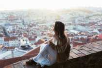 Vista lateral de la mujer joven en chaqueta y vestido sentado en la barrera de ladrillo y admirar paisaje urbano en la mañana soleada en Portugal - foto de stock