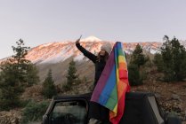 Piacevole donna scattare selfie su smartphone con bandiera LGBT seduta sul tetto dell'auto — Foto stock