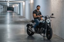 Ganzkörperbärtiger Typ in lässiger Kleidung, der auf einem Motorrad sitzt und im Flur einer modernen Garage in die Kamera blickt — Stockfoto