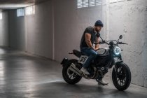 Corpo inteiro barbudo cara em roupas casuais sentado na motocicleta e olhando para longe no corredor da garagem moderna — Fotografia de Stock