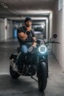 Ganzkörperbärtiger Typ in lässiger Kleidung, der auf einem Motorrad sitzt und im Flur einer modernen Garage in die Kamera blickt — Stockfoto