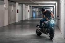 Full body bearded guy in casual clothes sitting on motorcycle and looking away in corridor of modern garage — Stock Photo