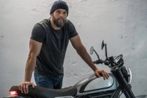 Full body bearded guy in casual clothes standing near on motorcycle and looking at camera in corridor of modern garage — Stock Photo