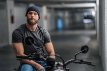 Corpo inteiro barbudo cara em roupas casuais sentado na motocicleta e olhando para a câmera no corredor da garagem moderna — Fotografia de Stock