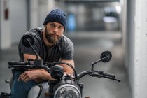 Full body bearded guy in casual clothes sitting on motorcycle and looking at camera in corridor of modern garage — Stock Photo