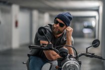 Full body bearded guy in casual clothes and sunglasses on motorcycle in corridor of modern garage — Stock Photo