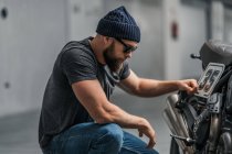 Bärtiger Mann mit Hut, der die Augenbraue hochzieht und in die Kamera blickt, während er in der Nähe von Motorrädern auf verschwommenem Hintergrund einer zeitgenössischen Garage sitzt — Stockfoto