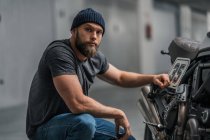 Bearded man in hat raising eyebrow and looking at camera while sitting near motorcycle on blurred background of contemporary garage — Stock Photo