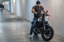 Повне тіло бородатого хлопця в повсякденному одязі, сидячи на мотоциклі і дивлячись в коридор сучасного гаража — стокове фото