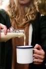 Корм женщины в теплой повседневной одежде заполнить белый металлический стакан с вкусным молочным напитком из стеклянной бутылки во время пикника на природе — стоковое фото