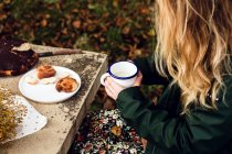 From above crop female in warm casual clothes drinking milk and eating delicious homemade pastry while sitting at gray stone table with bouquet of fresh yellow wildflowers in autumn garden — Stock Photo