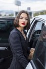 Gorgeous young brunette with red lipstick in stylish outfit looking at camera while getting into cat at city street — Stock Photo