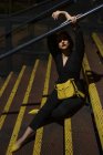 Mujer de moda en vestido negro con lápiz labial rojo y bolsa pequeña amarilla que se mete en la tapa de pico sentado en la barandilla de la escalera en una calle de la ciudad en el atardecer - foto de stock