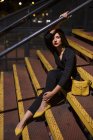 Модна жінка в чорній сукні з червоною помадою та жовтим мішком, що сидить на поручнях сходів на вулиці міста в сутінках — стокове фото