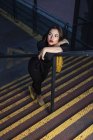 Femme à la mode en robe noire avec rouge à lèvres rouge et petit sac jaune penché à la main courante de l'escalier dans une rue de la ville au crépuscule — Photo de stock