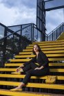 Mulher na moda em vestido preto com batom vermelho e pequeno saco amarelo sentado no corrimão da escada em uma rua da cidade no crepúsculo — Fotografia de Stock